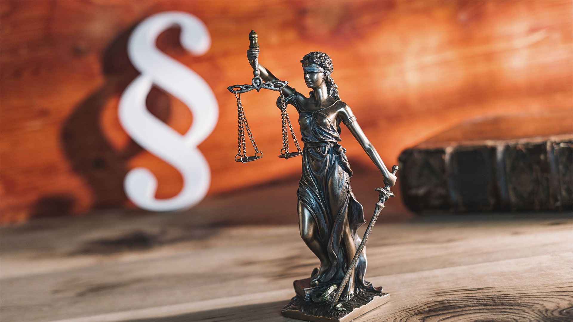 Auf einer hölzernen Oberfläche steht eine Justitia Figur, im Hintergrund ist ein Paragrafen-Symbol zu sehen